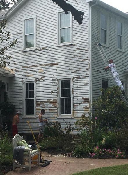 People painting a two-story white house; paint peeling from the siding.
