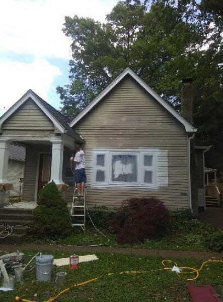 Person painting white trim on the side of a house with green siding, standing on a ladder.