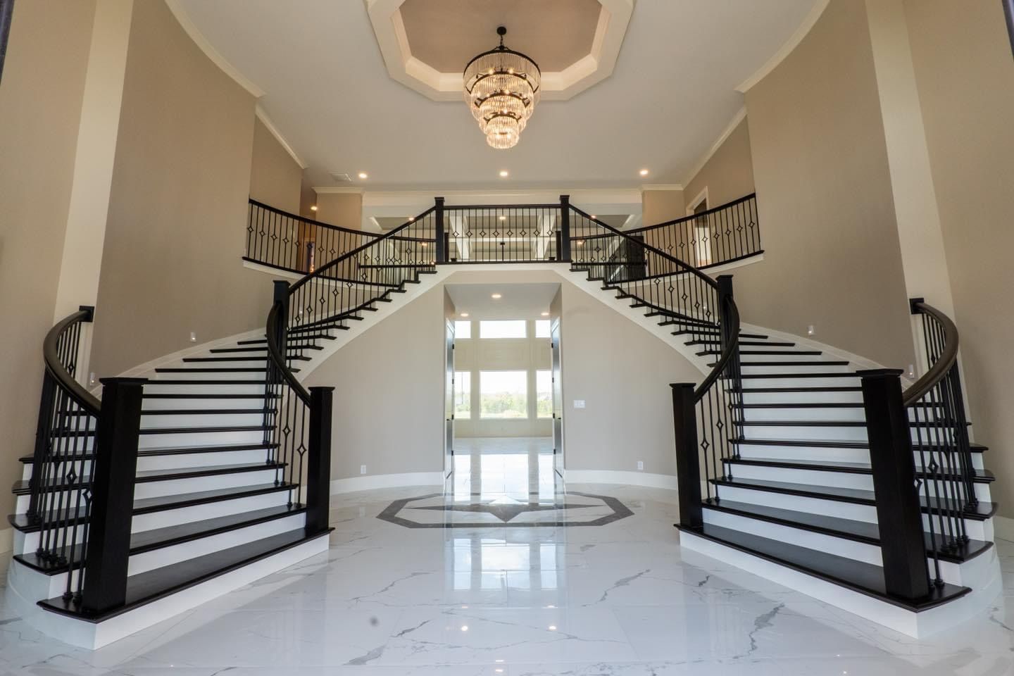 Grand foyer with dual staircases, chandelier, and white marble floor. Black banisters and beige walls.