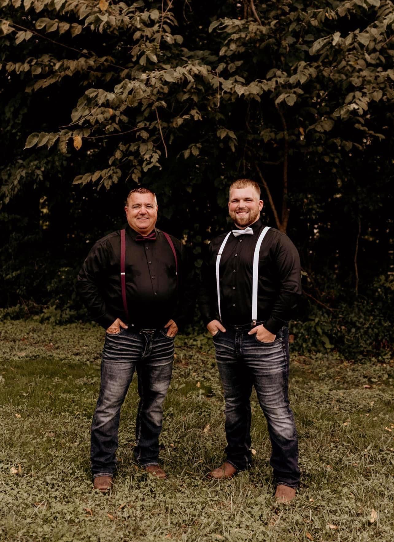 Two men in suspenders and jeans stand outside; one with a red bow tie, the other with a white bow tie.