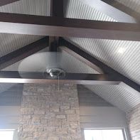 Beamed ceiling with fan, brick fireplace, corrugated metal roof, and windows.