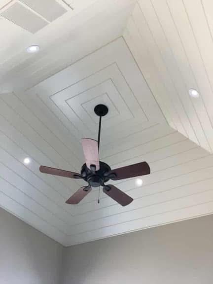 Ceiling with a dark ceiling fan, recessed lights, and layered square design. Wooden paneling is painted white.