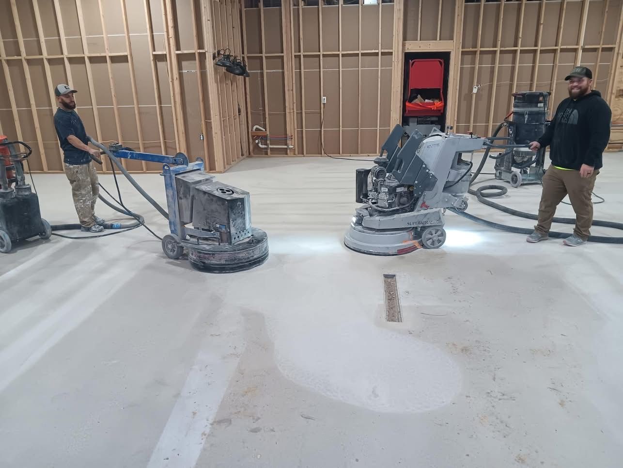 Two men grinding a concrete floor with industrial machines in an unfinished building.