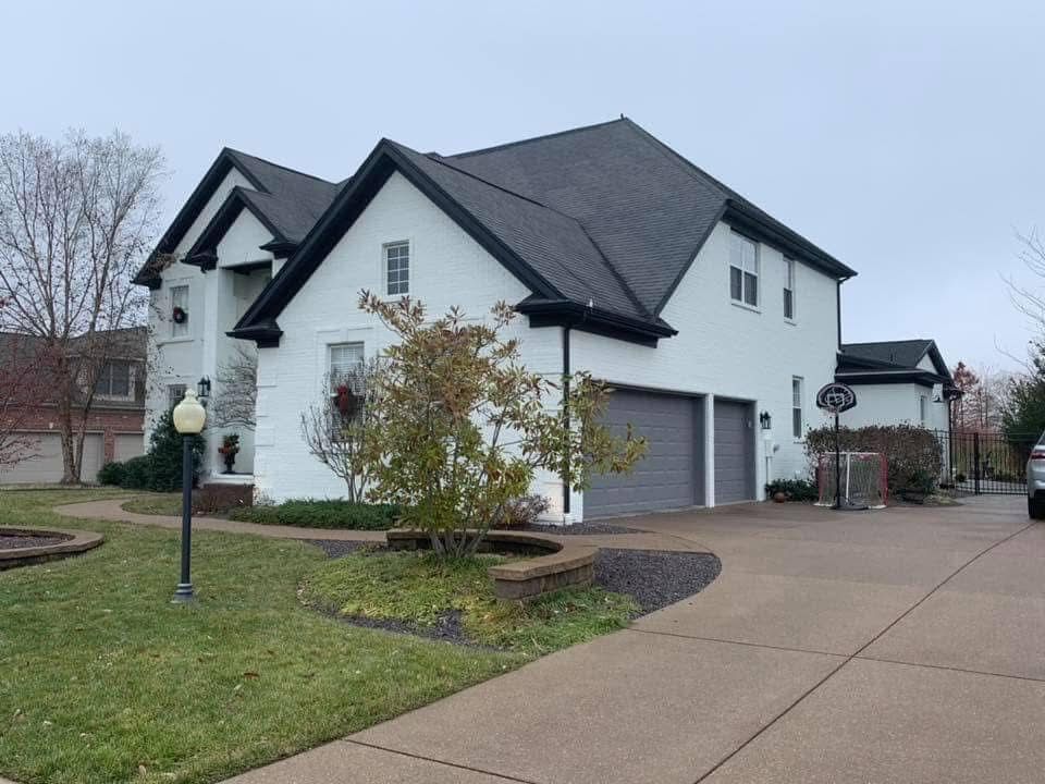 White two-story house with black trim and a gray driveway. A small tree sits in front. Overcast sky.