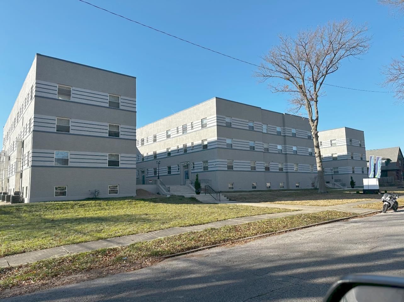 Three-story apartment buildings with light gray facades and small windows under a clear, blue sky. A bare tree stands nearby.