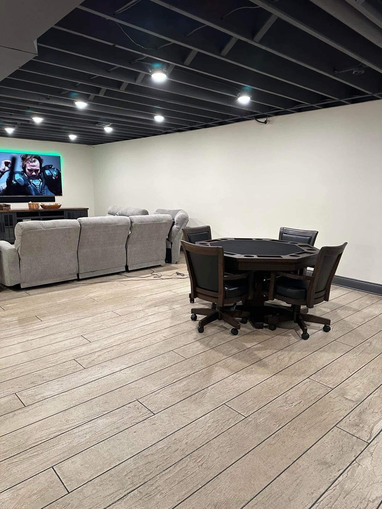 Basement recreation room with a sectional sofa, TV, and game table on wood-look tile flooring.