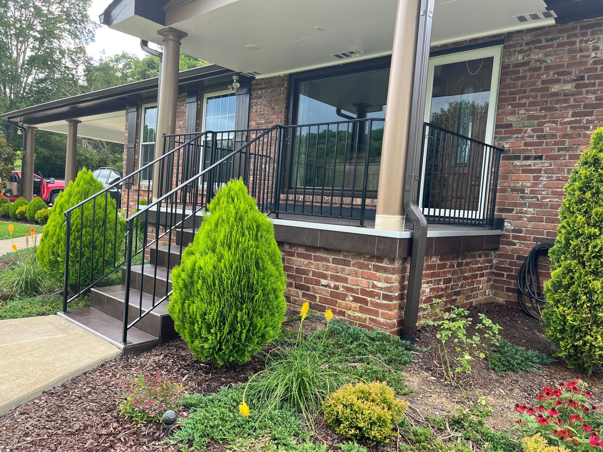 A brick house with a porch and stairs surrounded by bushes and flowers.