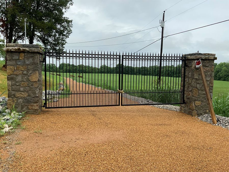 A wrought iron gate surrounds a gravel road leading to a field.
