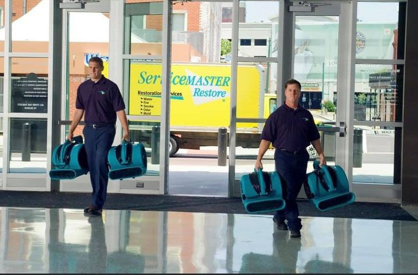 Two ServiceMaster employees in uniform carrying teal equipment bags, exiting a building with a yellow service truck in the background.