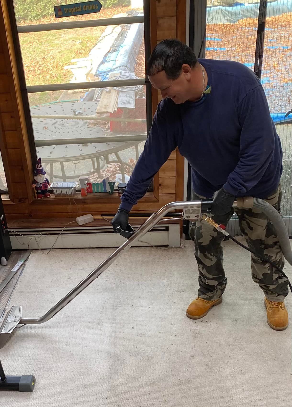 Man in camo pants cleaning carpet with a machine near a window.