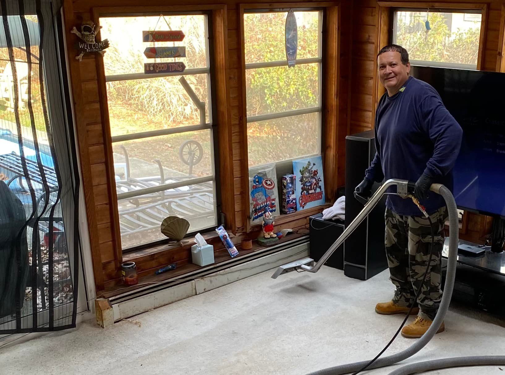 Man cleaning a beige carpet with a vacuum in a room with windows. He wears gloves and camo pants.