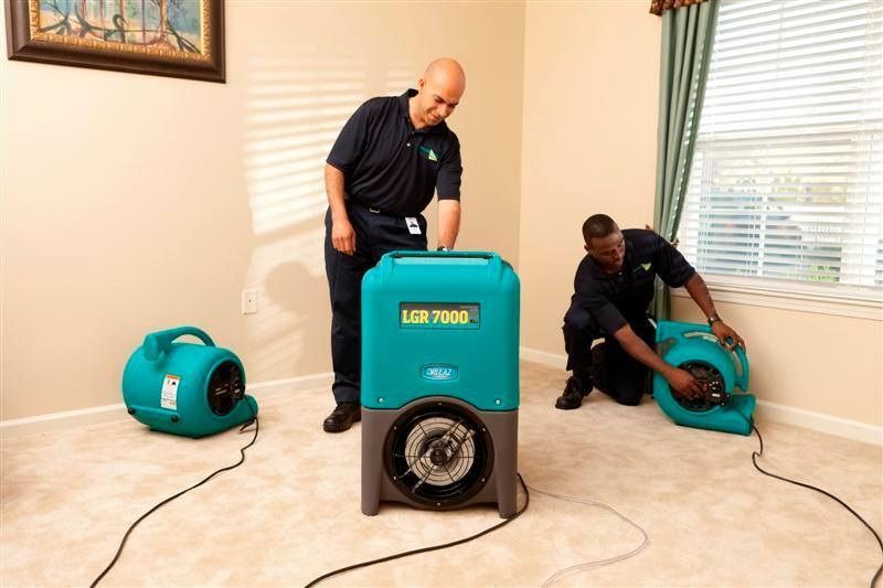 Two men setting up air drying equipment in a carpeted room. Teal and black equipment.