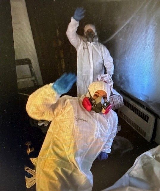 Two people in protective suits wave inside a room with plastic sheeting.