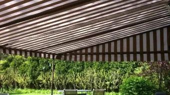 Shaded patio with striped roof, green hedge, and bench overlooking lush garden