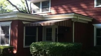 Red house porch with striped awning and white-trimmed windows, partly shaded by bushes