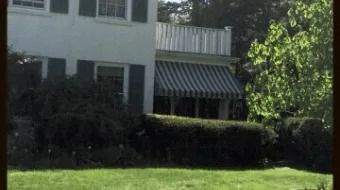 White house with striped awning, shaded hedge, and green lawn in front.