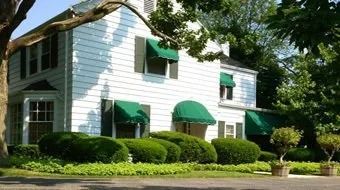 White two-story house with green awnings and shrubs, viewed from the street