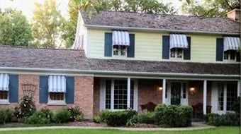Two-story brick and siding house with blue shutters, white awnings, and a front porch surrounded by shrubs