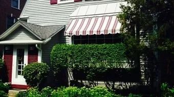 House exterior with red-and-white striped awning, red shutters, and neatly trimmed green hedges.