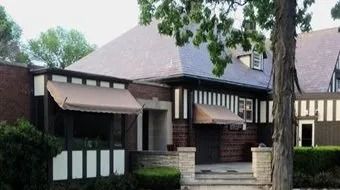 Brick house with striped awnings, front steps, and manicured shrubs under trees