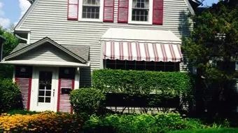 White house with red shutters, striped awning, and front porch surrounded by lush greenery.