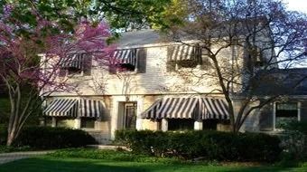 Two-story cream house with striped awnings, surrounded by trees and shrubs