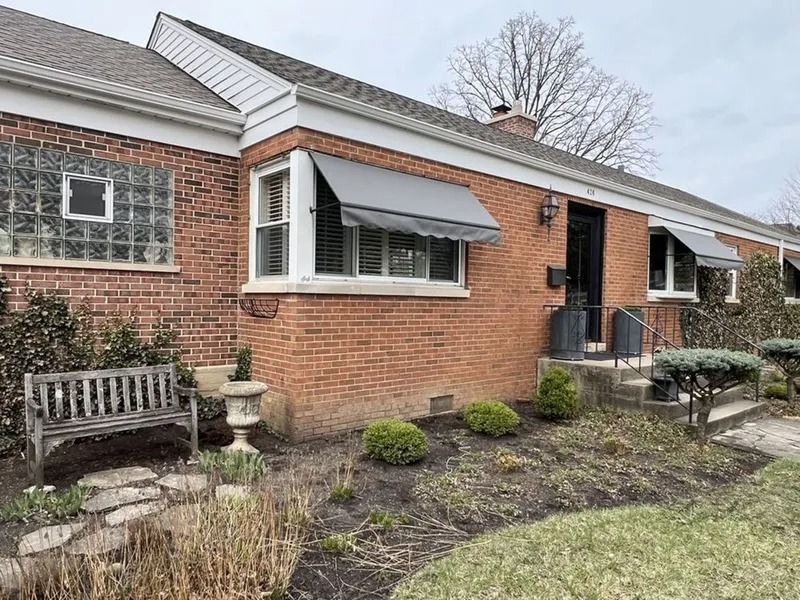 Brick house exterior with white-trimmed bay window, awning, bench, and small landscaped garden