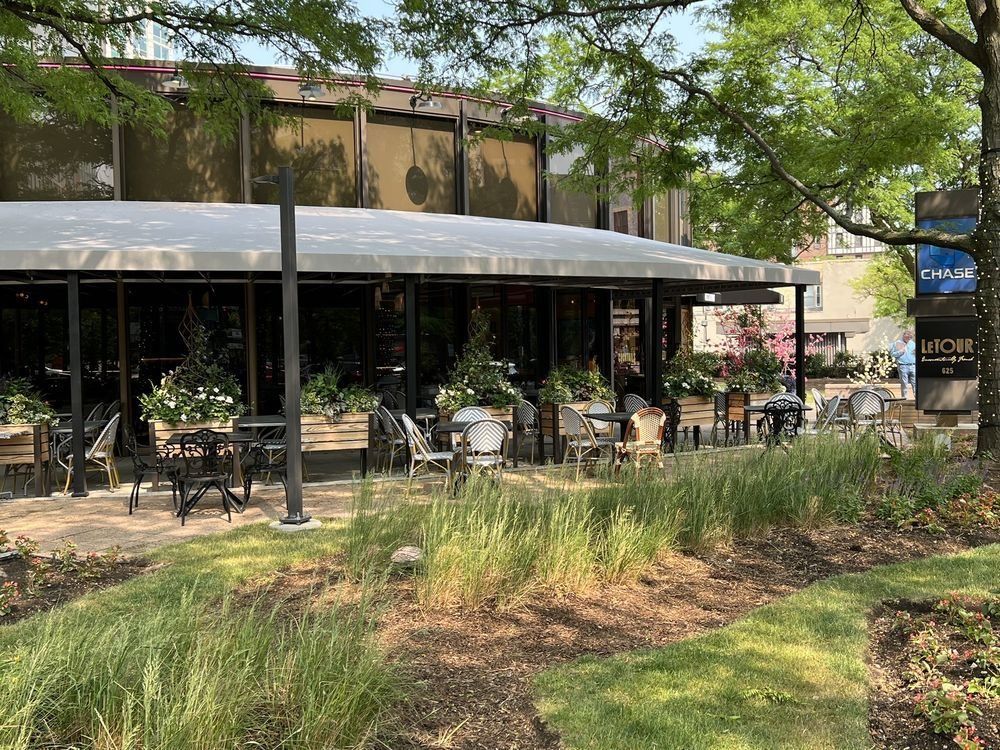 Outdoor café patio with black umbrellas, tables, and chairs beside a landscaped garden under trees