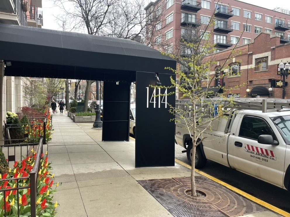 Apartment entrance with black awning, sidewalk planters, and parked SUV on a city street