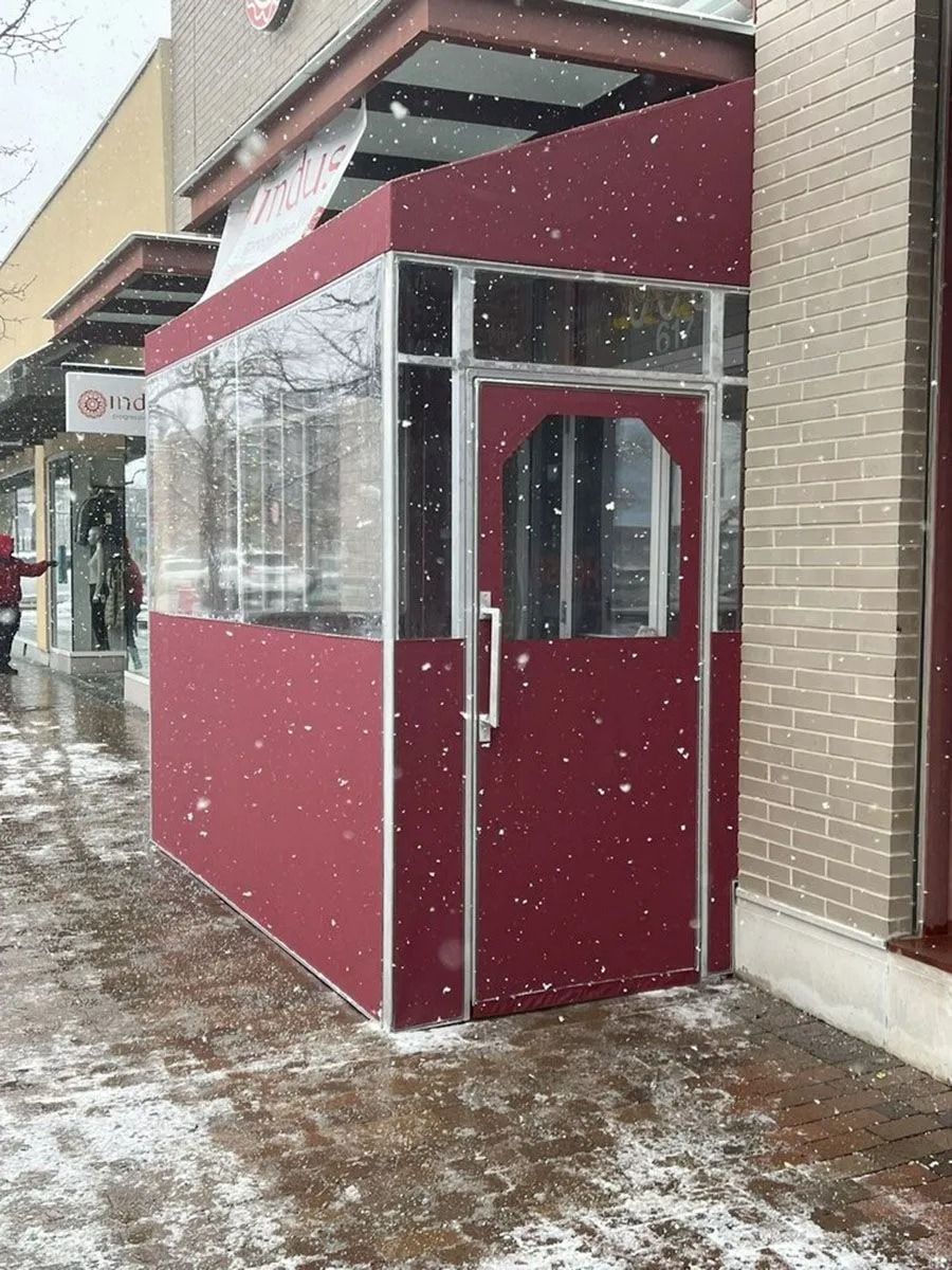Maroon outdoor kiosk with clear windows and a slanted awning on a snowy sidewalk.