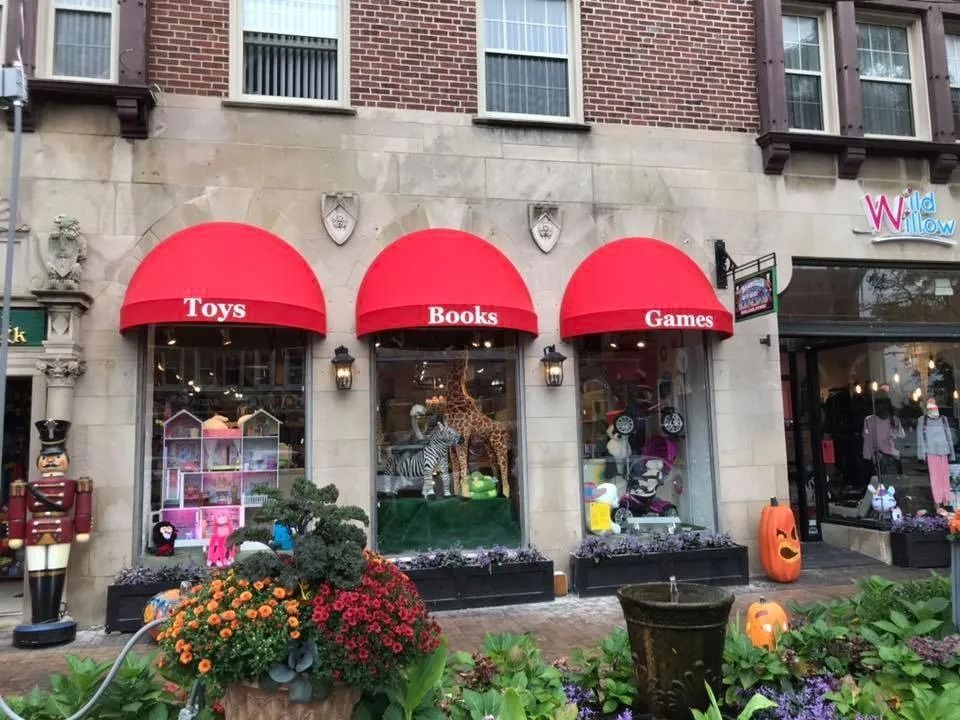 Storefront with red awnings labeled Toys, Books, and Games, with flowers and Halloween pumpkins outside