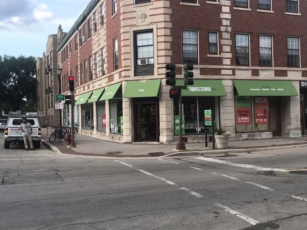 Corner storefronts with green awnings on a city street intersection, with a pedestrian and parked car.