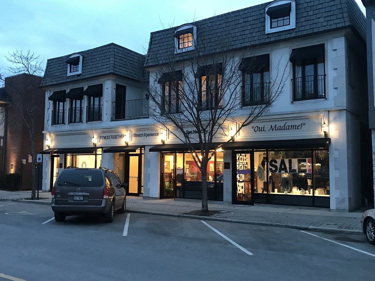 Two-story storefronts at dusk with lit windows and a parking lot, one shop displaying a large SALE sign.