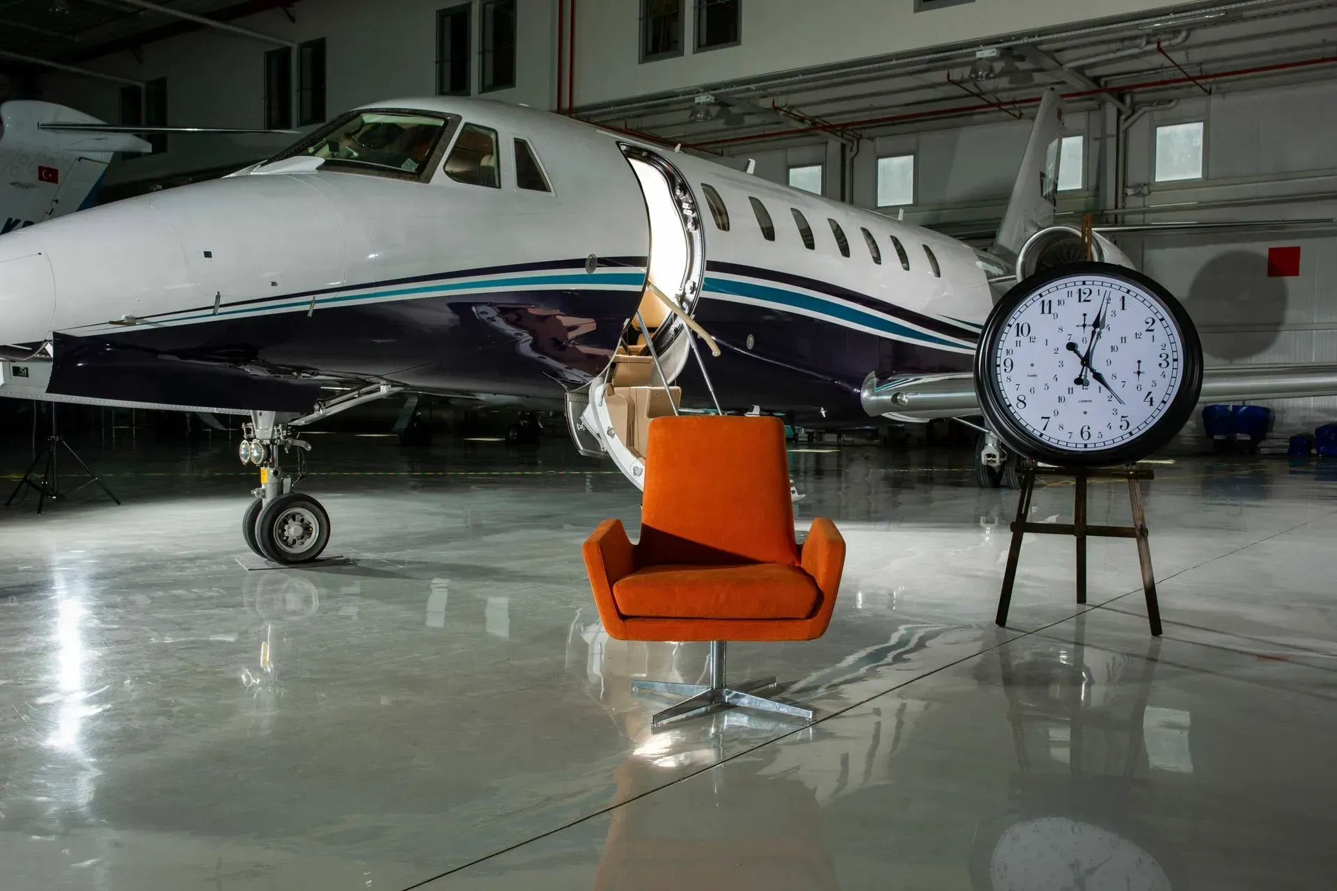 Jet airplane with open door, orange chair, and large clock in a hangar.