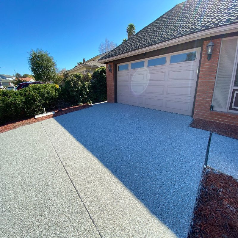 Driveway with a garage door and house exterior. Driveway is light gray, with brown mulch and greenery. Blue sky.