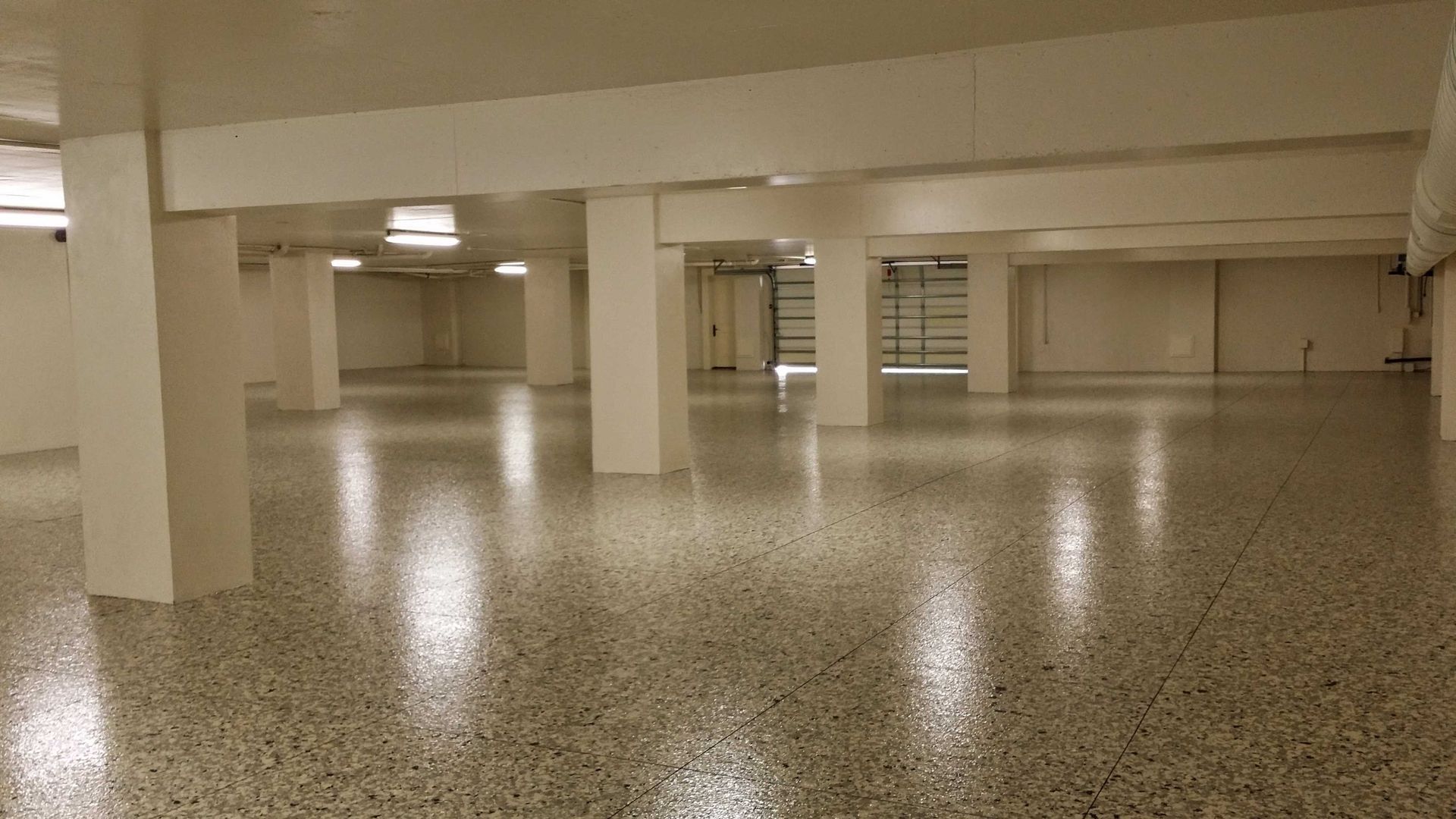Empty, brightly lit, beige-toned room with polished speckled floor, support columns, and recessed lighting.