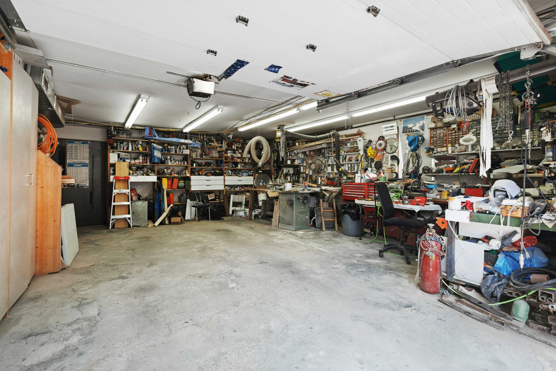 Cluttered garage interior, various tools and equipment, workbench, overhead lights, concrete floor.