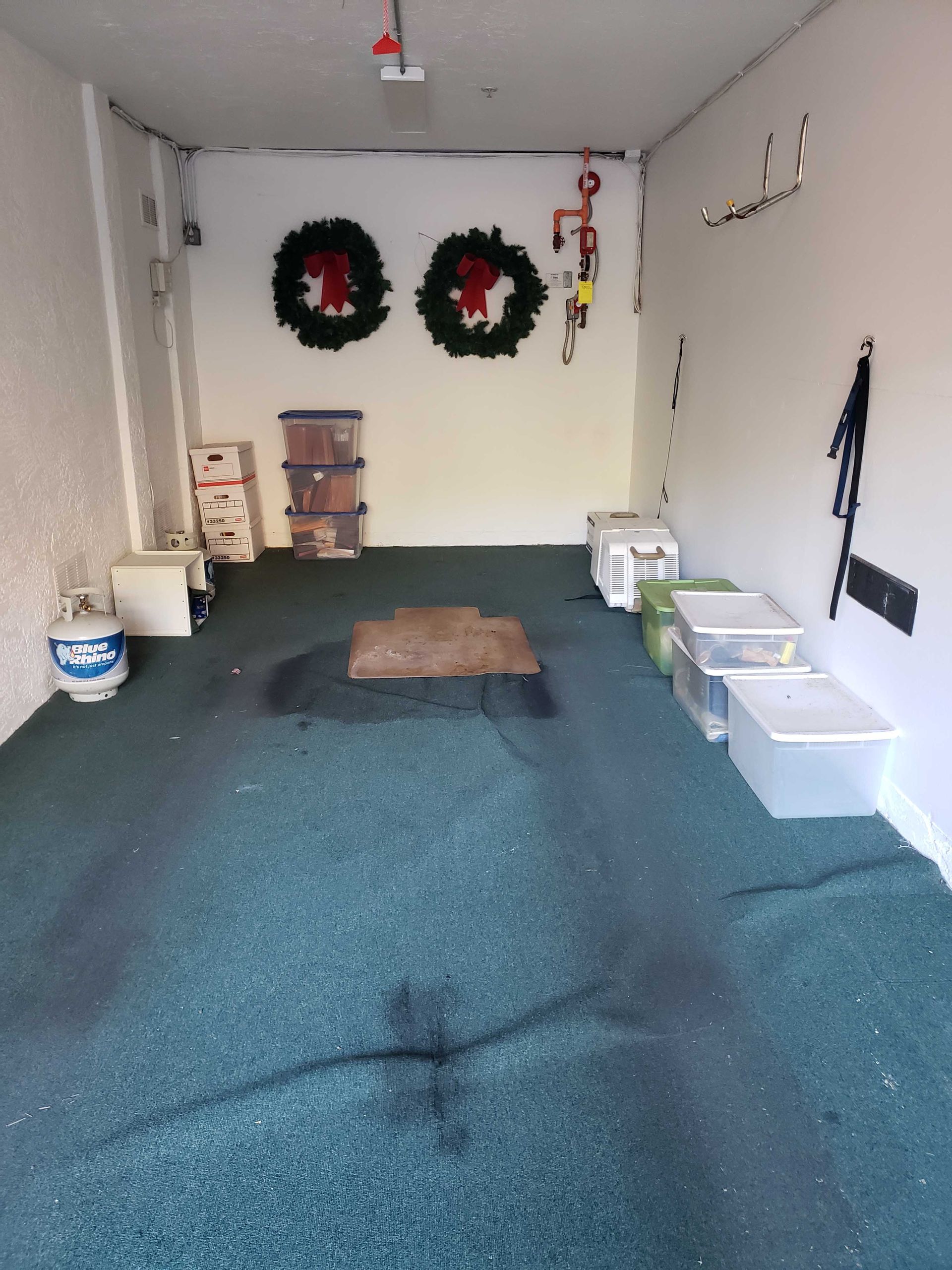A cluttered garage with green floor, storage containers, propane tank, and Christmas wreaths.
