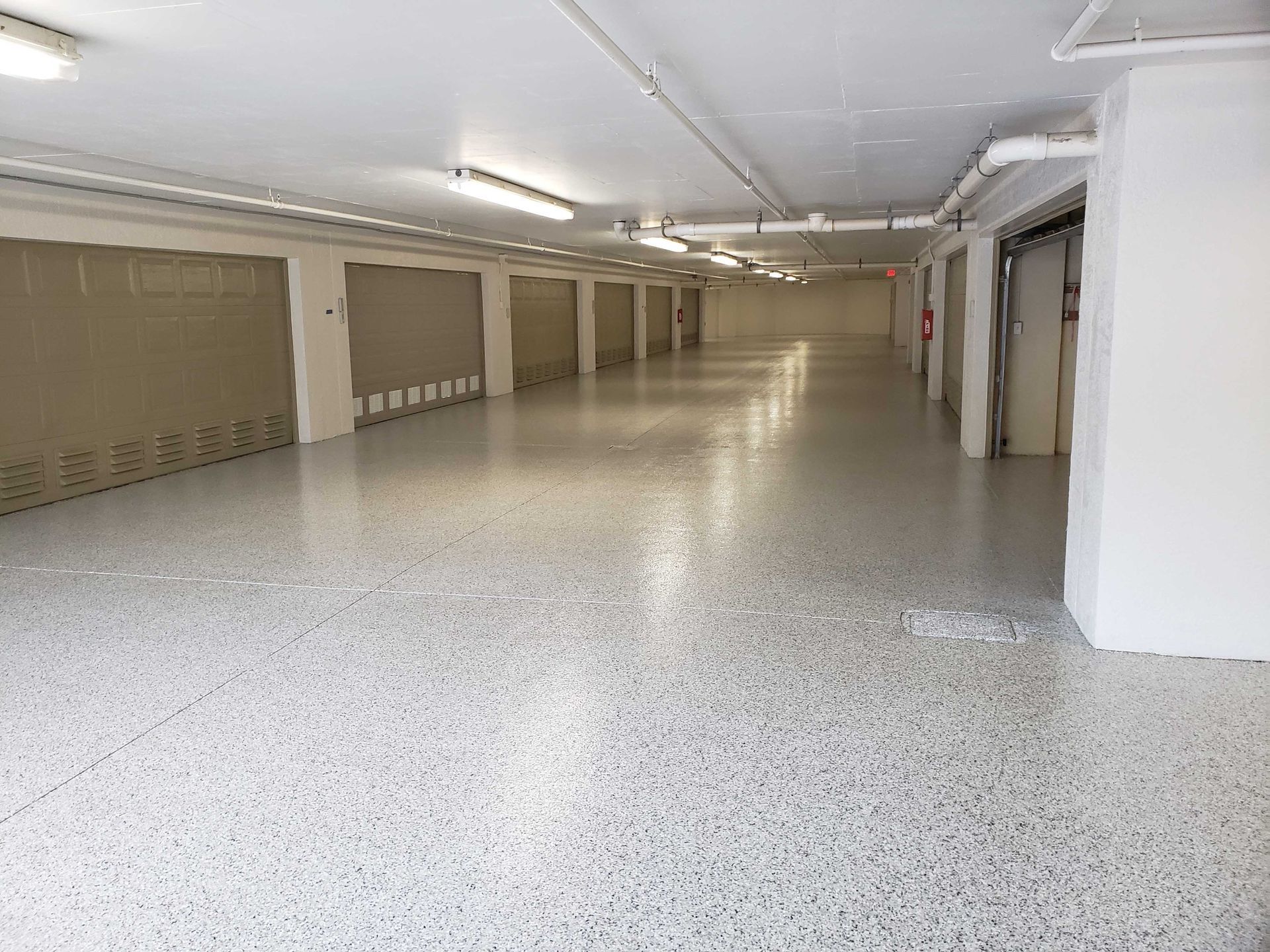 Brightly lit storage unit hallway with gray speckled flooring and closed tan roller doors.