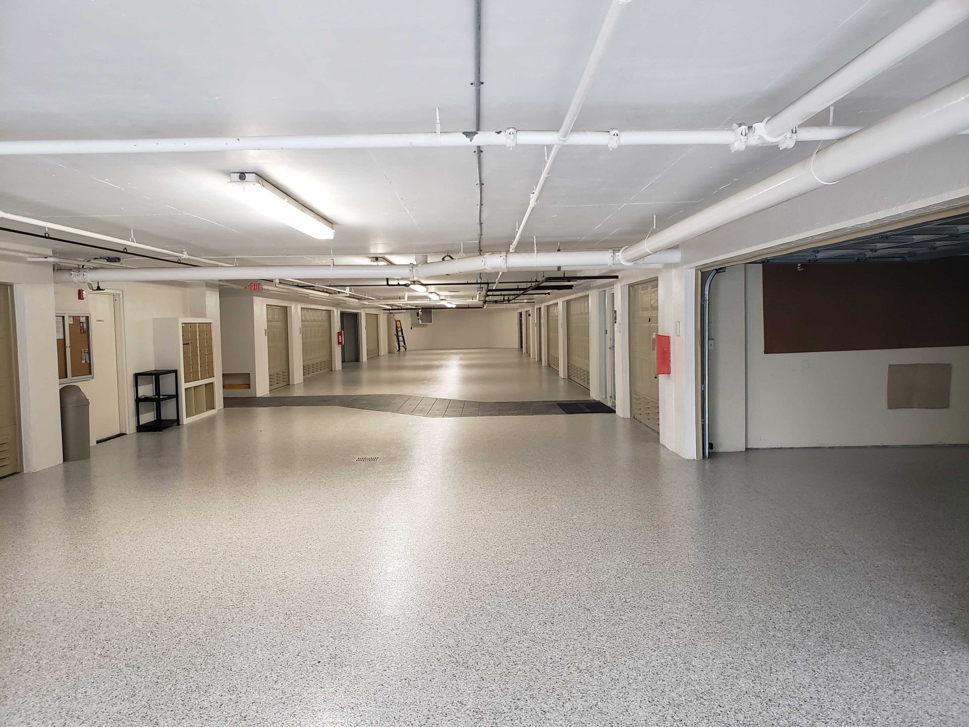 A long, empty garage with rows of storage units, white walls, speckled floor, and overhead lighting.