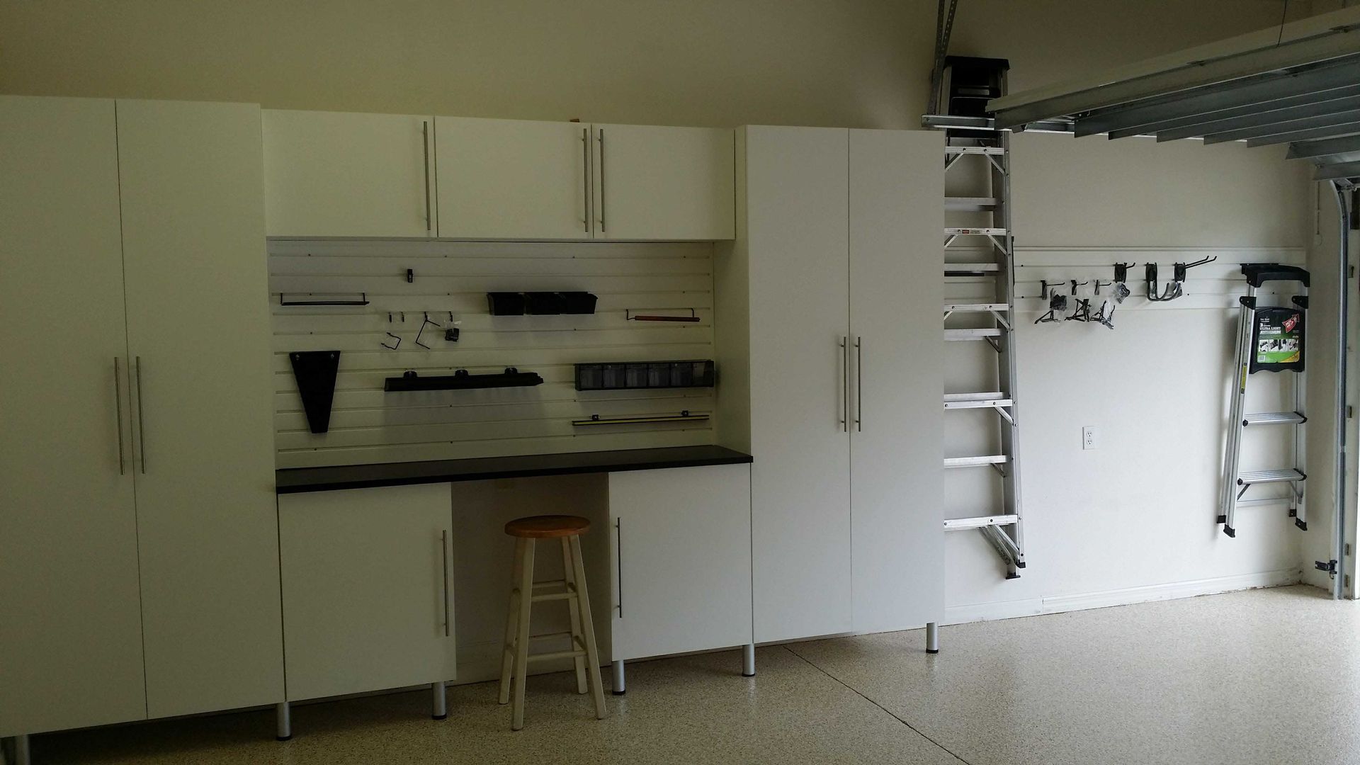 White garage cabinets with a workbench and pegboard holding tools. A stool sits beneath the workbench.