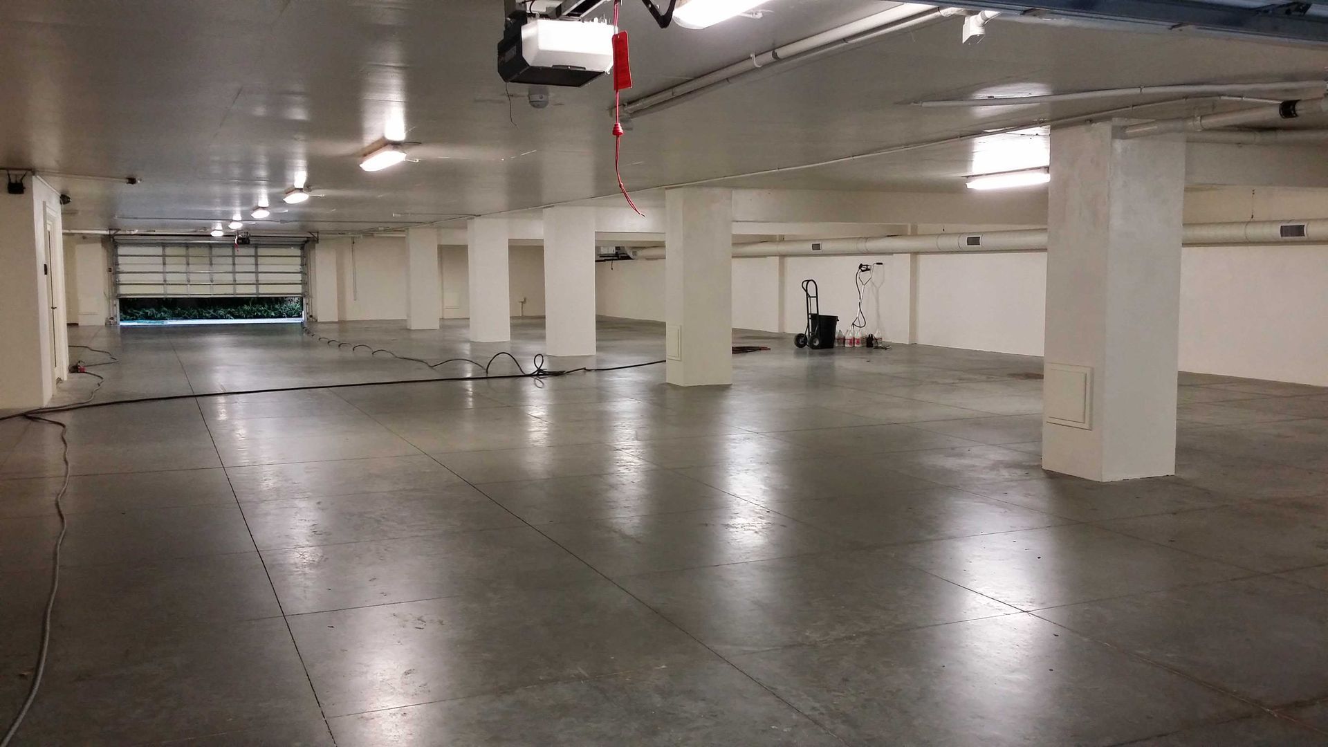 Empty, brightly lit, concrete-floored parking garage. White columns support the ceiling. Garage door open at far end.