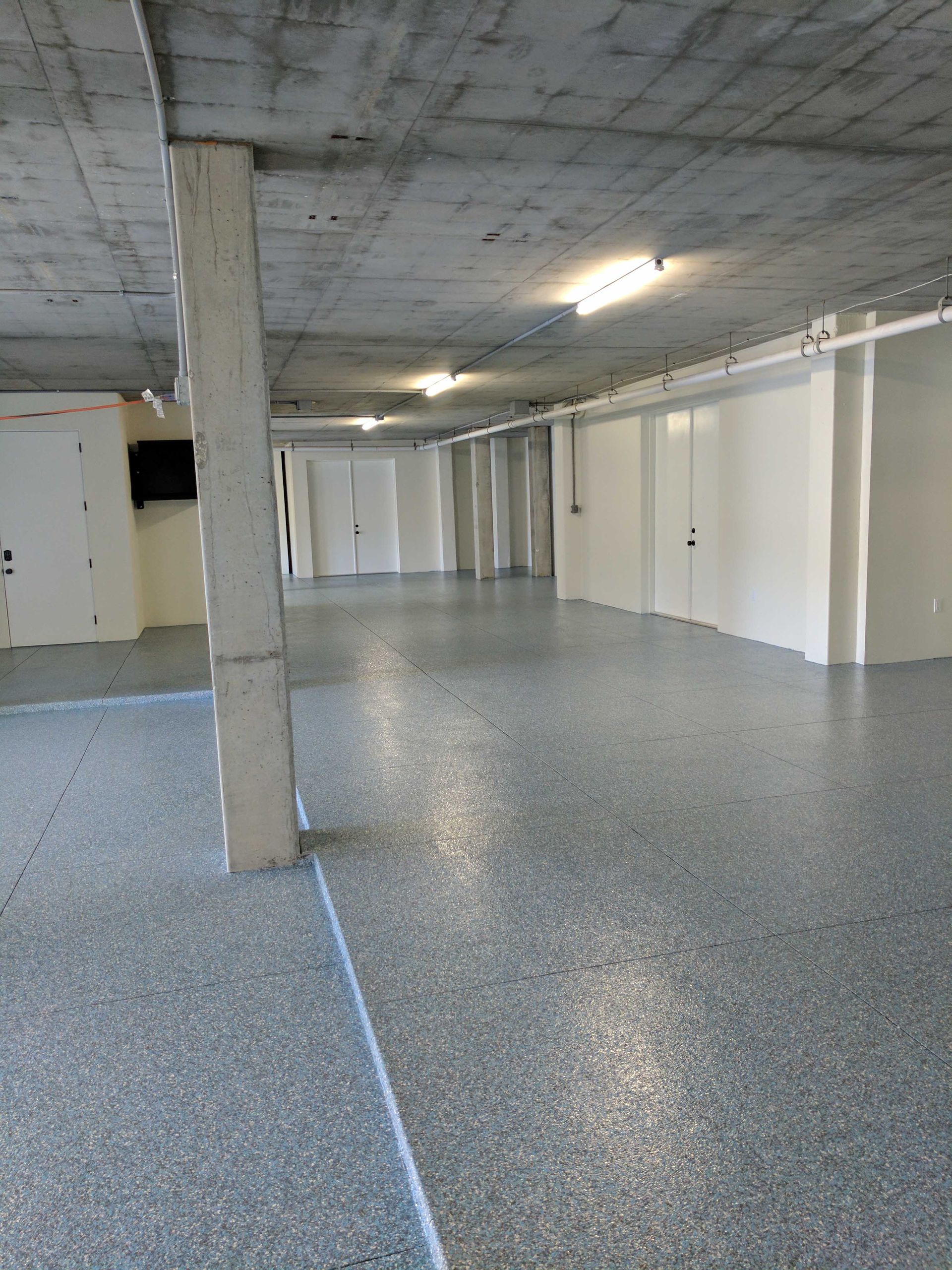 Concrete-floored garage/storage space; grey and white; several white storage units/doors line the walls.