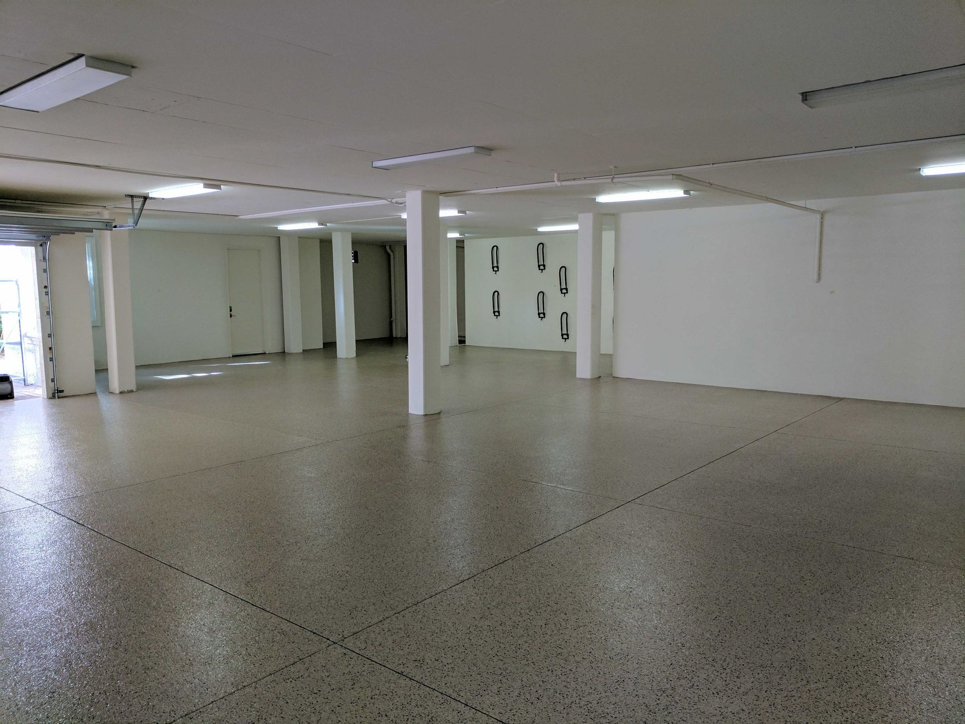 Empty white-walled gallery space with speckled flooring, illuminated by overhead lights.