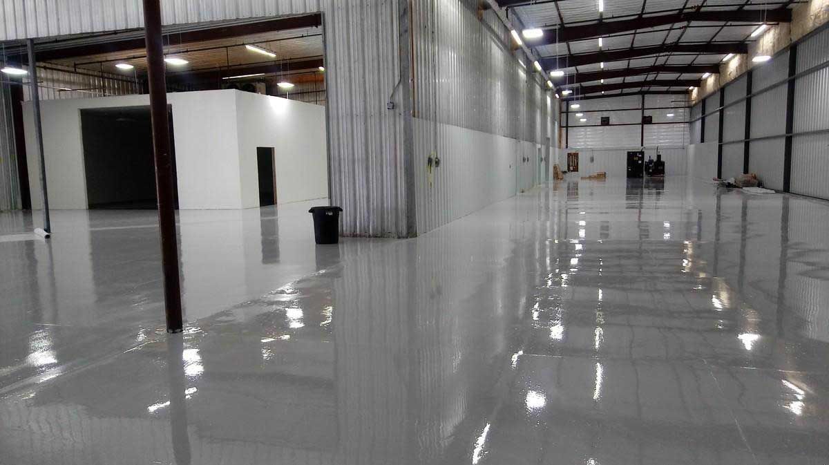 A shiny, light gray epoxy floor in a large, empty warehouse with reflective surfaces.