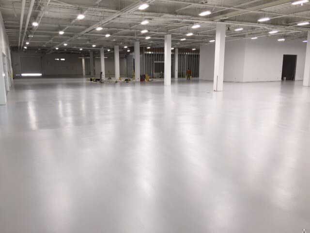 Large, empty room with shiny grey floor and white pillars. Rows of overhead lights.