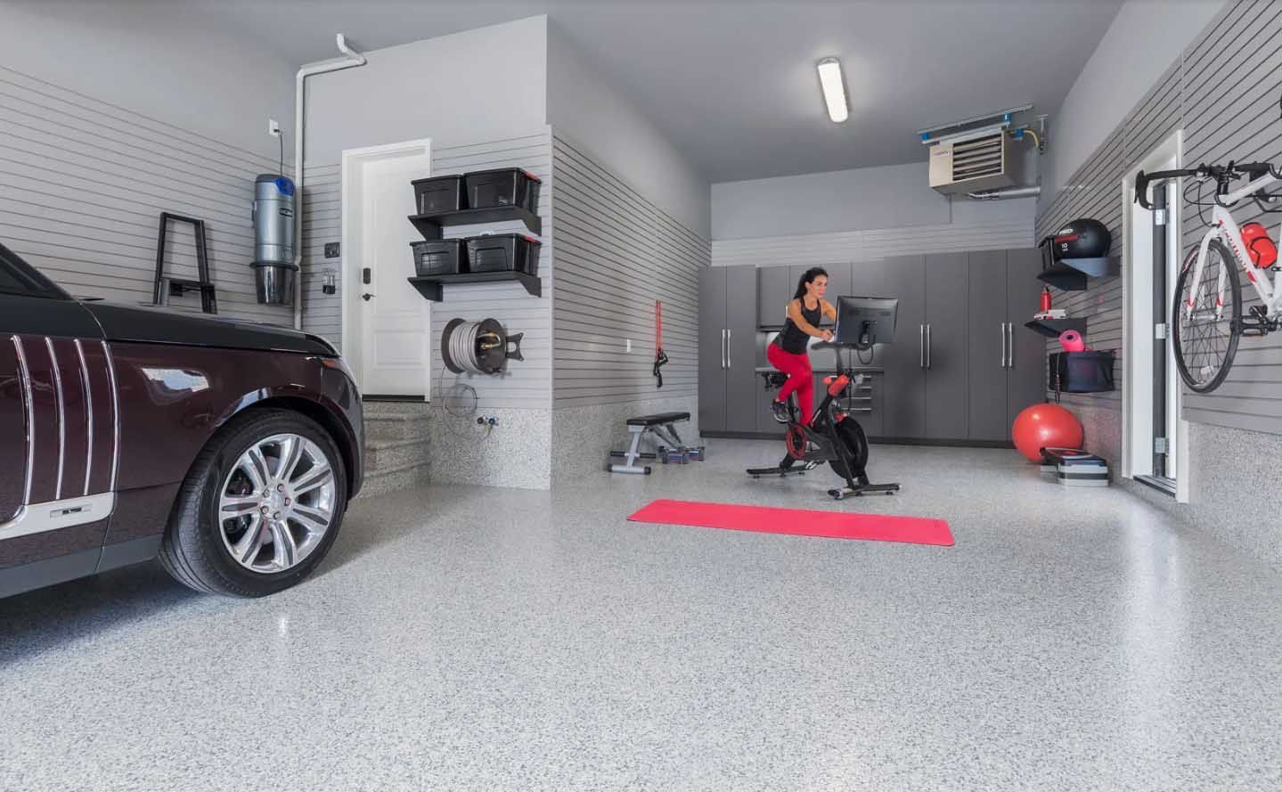 Woman exercises in a garage gym. Car, bike, equipment, and gray speckled floor.