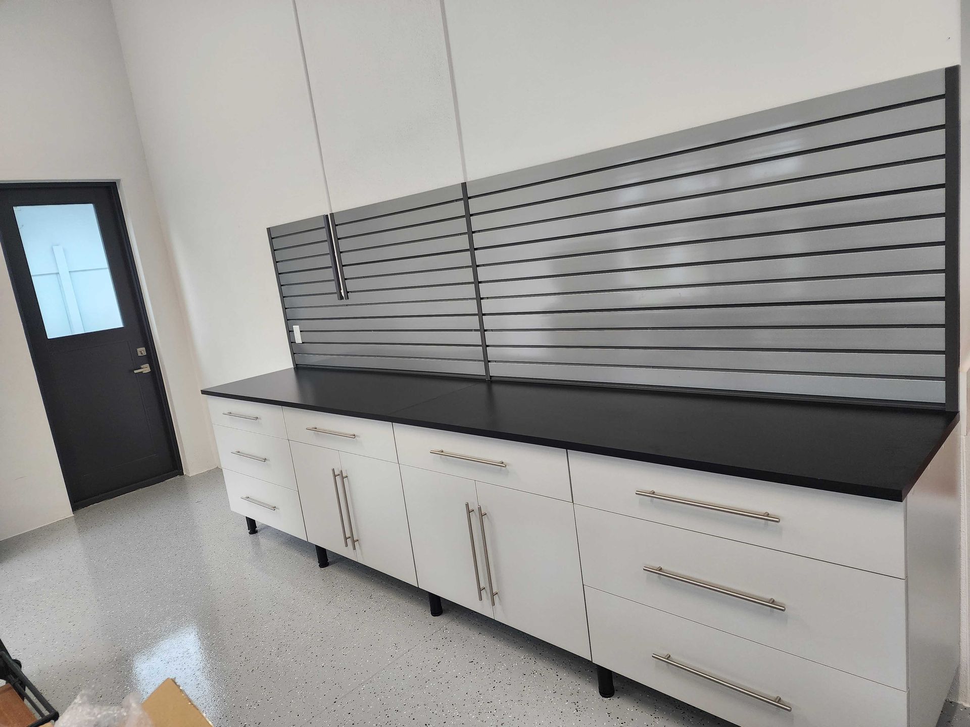 Garage with white cabinets, black countertop, and grey wall organizer. Dark door on the left.