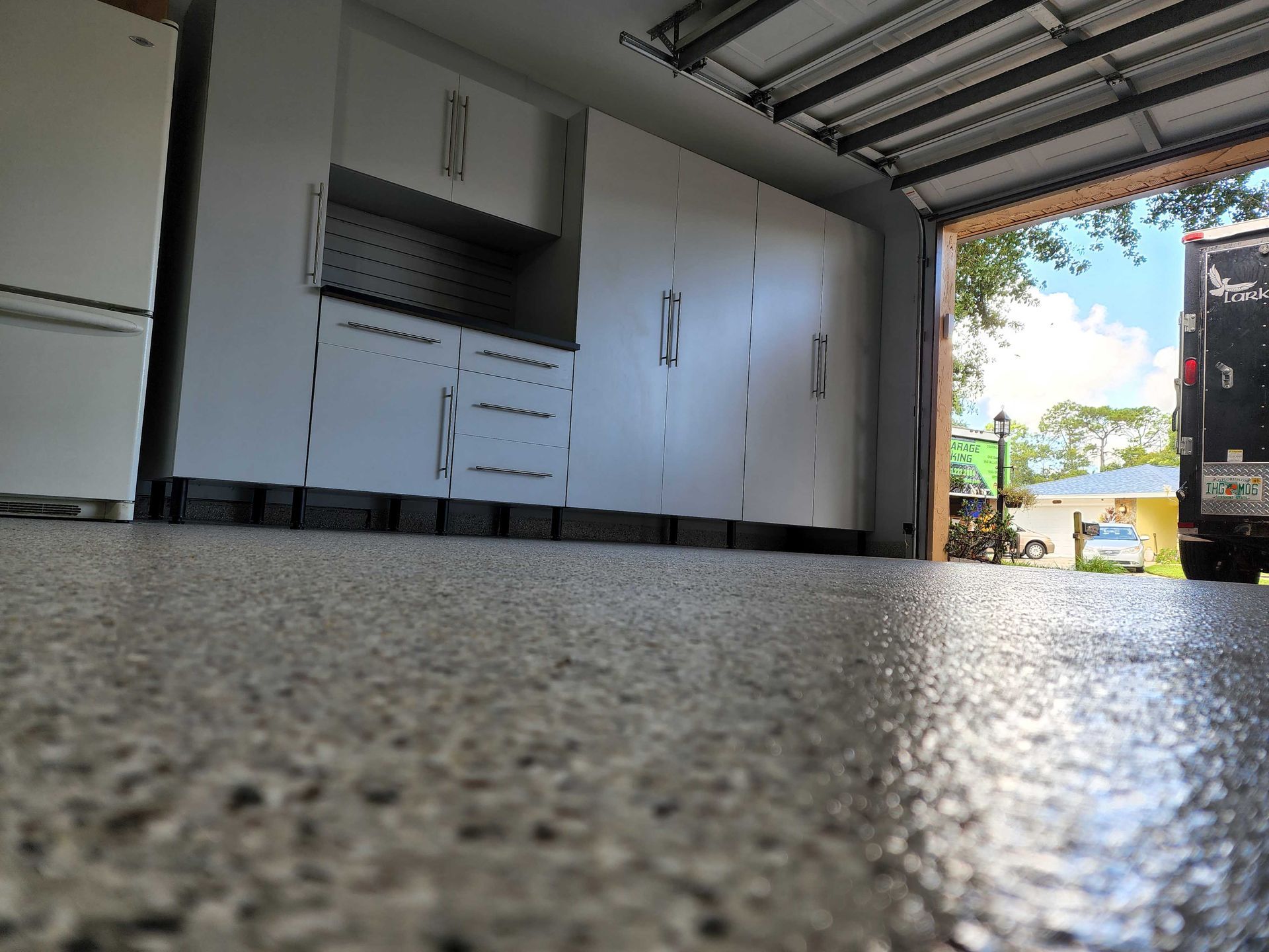Low-angle view of a garage with white cabinets and a gray epoxy floor. The garage door is open, with a vehicle in the distance.