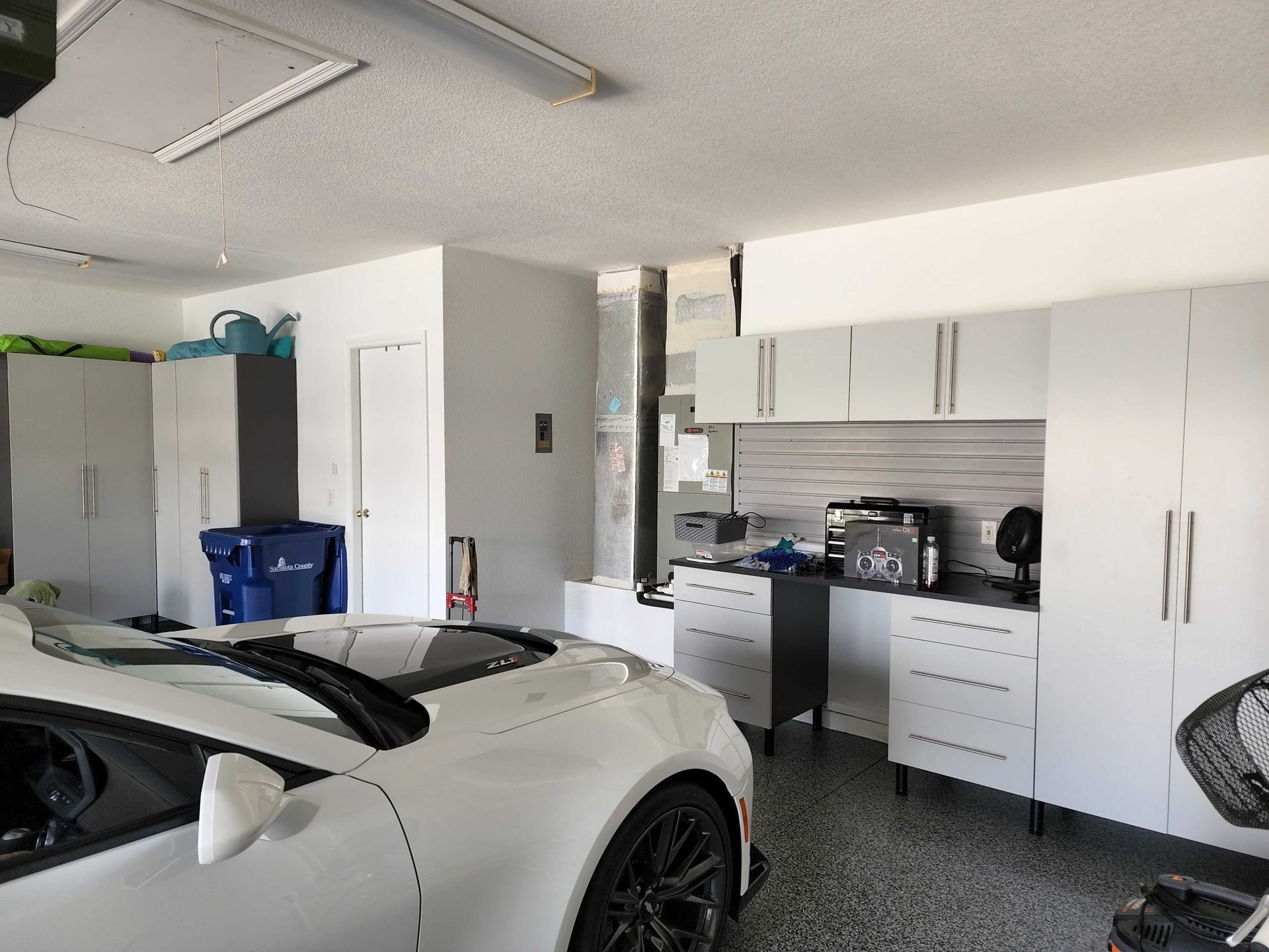 White sports car in a garage with gray cabinets, appliances, and speckled floor.